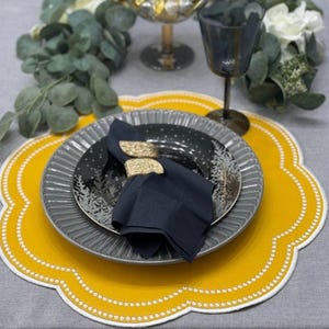 May include: A table setting featuring a yellow placemat with a scalloped edge and white dot trim. A gray plate with a black and silver plate on top, a black napkin, and a gold napkin ring complete the elegant look.
