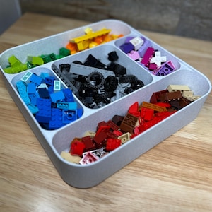 May include: A gray plastic organizer filled with colorful toy building blocks. The blocks are sorted into different compartments, including blue, green, yellow, red, black, and purple pieces. The organizer sits on a wooden surface.