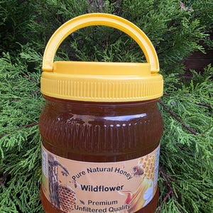 Peut inclure: Un pot en plastique transparent rempli de miel de fleurs sauvages ambré, doté d'un couvercle à vis jaune vif et d'une poignée jaune assortie. L'étiquette porte le texte Pure Natural Honey, Wildflower, et Premium Unfiltered Quality.