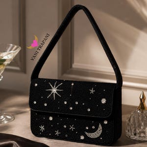 May include: Black beaded handbag with a flap closure, adorned with a celestial design of stars, a crescent moon, and sparkling crystals. The bag has a matching beaded handle and the text "VANI TRAPANI".