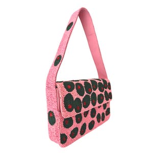 May include: A pink beaded shoulder bag with a flap closure. The bag features a pattern of dark green and red circular designs. The strap and the body of the bag are covered in small pink beads. The bag is shown against a white background.