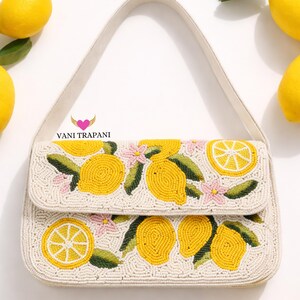 May include: Cream-colored beaded handbag with a flap closure, featuring a lemon design. The design includes yellow lemons, green leaves, and pink flowers. The bag has a white strap and the text "Vani Trapani".
