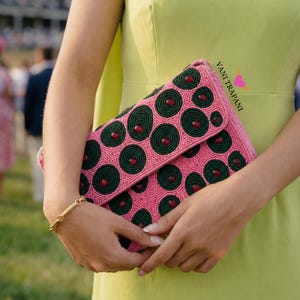 May include: Pink beaded clutch with a flap closure. The clutch features a pattern of dark green circles with red accents. A gold bracelet is visible on the wrist. The text "Vani Trapani" is printed on the clutch.