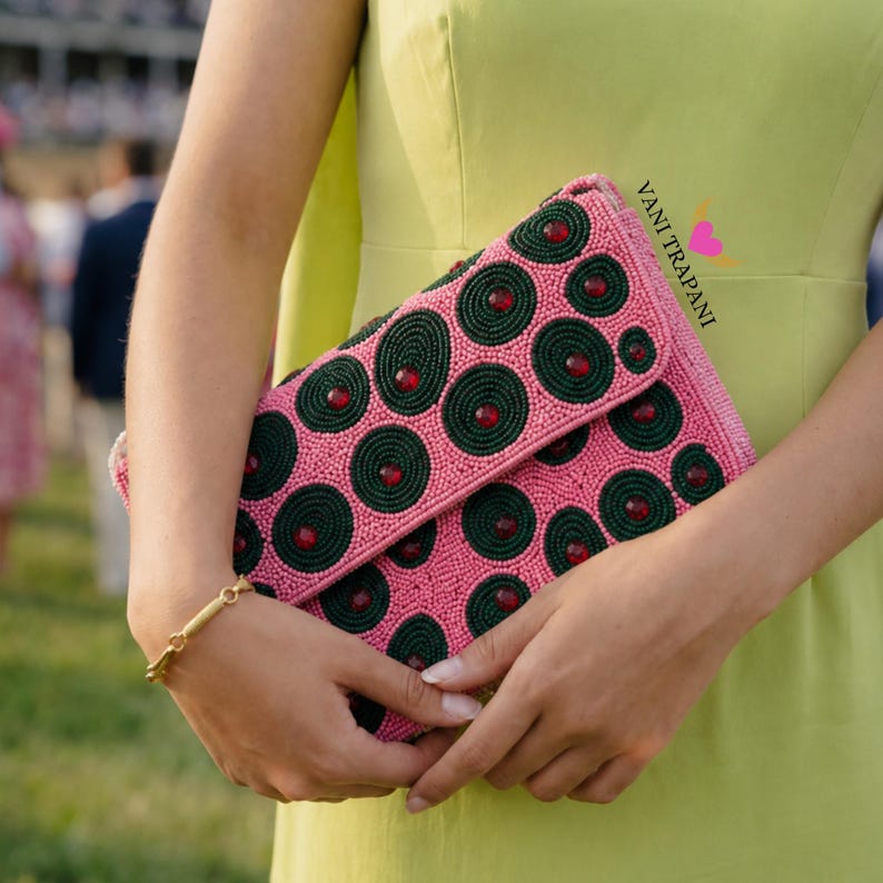 May include: Pink beaded clutch with a flap closure. The clutch features a pattern of dark green circles with red accents. A gold bracelet is visible on the wrist. The text "Vani Trapani" is printed on the clutch.