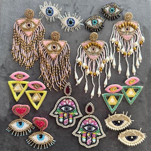May include: A selection of beaded earrings in diverse designs. Styles include eye-shaped, triangle, and Hamsa hand shapes, with pink, gold, blue, and white beads. Some earrings have dangling fringe.
