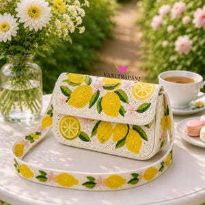 May include: A white handbag with a flap closure, adorned with embroidered yellow lemons, green leaves, and pink flowers. The matching strap also features the lemon design. The text "VANI TRAPANI" is visible.
