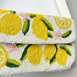 May include: White beaded clutch with a lemon design. The clutch features yellow lemons, green leaves, and pink flowers. The strap is white.