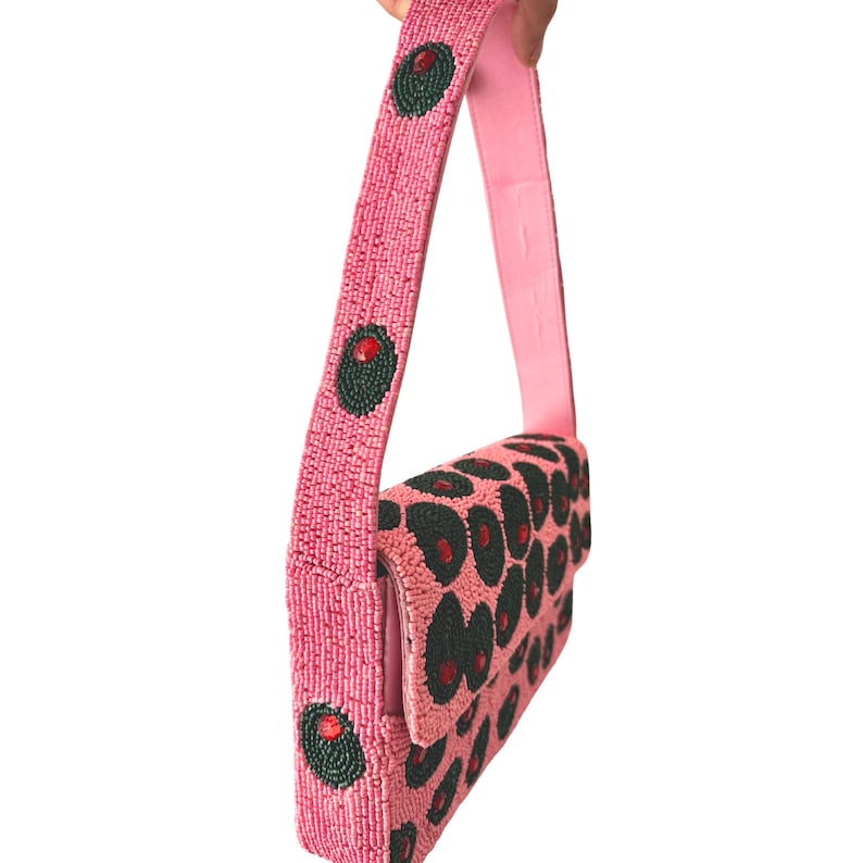 May include: Pink beaded handbag with a flap closure and a shoulder strap. The bag features a pattern of dark green and red circular designs. The strap has green and red circular designs. The bag is held against a white background.