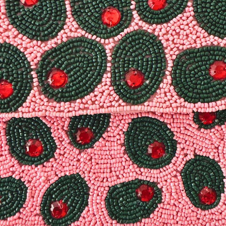 May include: Close-up of a beaded clutch with a pink background. The clutch features dark green beaded oval shapes with red rhinestone centers. The background is covered in small pink beads.