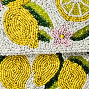 May include: Close-up of a beaded clutch with a lemon design. The clutch features yellow lemons, white and yellow lemon slices, green leaves, and pink flowers, all against a white background. The beads are small and tightly packed.