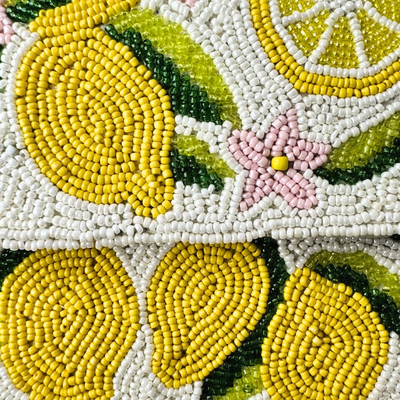 May include: Close-up of a beaded clutch with a lemon design. The clutch features yellow lemons, white and yellow lemon slices, green leaves, and pink flowers, all against a white background. The beads are small and tightly packed.