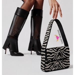 May include: A pair of black knee-high boots featuring sheer mesh panels and pointed toes, paired with a rectangular shoulder bag covered in a black and silver zebra-striped beaded pattern. The bag features the text VANI TRAPANI with a heart logo.