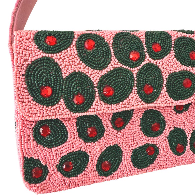 May include: Pink beaded handbag with a flap closure and a pink strap. The bag is covered in dark green beaded circles with red beaded accents, resembling olives. The bag is a unique accessory.