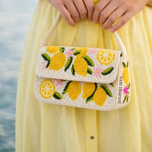 May include: Off-white beaded handbag with a flap closure, featuring a lemon design. The bag has yellow lemons, green leaves, and pink flowers. The strap and the body of the bag are off-white. The text "VANI TRAPANI" is visible on the side.