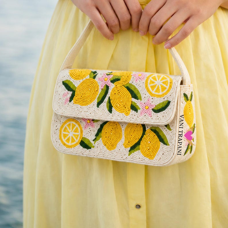 May include: Off-white beaded handbag with a flap closure, featuring a lemon design. The bag has yellow lemons, green leaves, and pink flowers. The strap and the body of the bag are off-white. The text "VANI TRAPANI" is visible on the side.