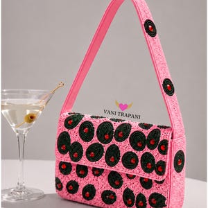May include: Pink beaded handbag with a flap closure, adorned with dark green and red circular patterns. The bag has a matching pink handle and the text "VANI TRAPANI". A martini glass with an olive is in the background.