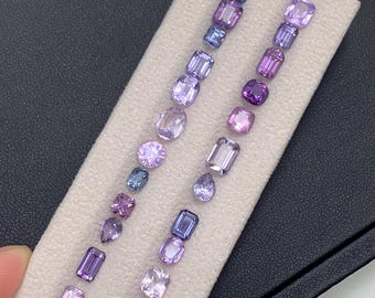 May include: An assortment of purple gemstones in various shapes and sizes, including emerald cuts, ovals, and pear shapes. The gems are arranged on a light beige fabric, showcasing a range of violet hues from light lavender to deep amethyst.