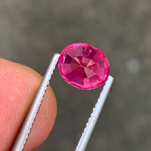 May include: A faceted, oval-cut, hot pink gemstone is held by silver tweezers. The gem has a brilliant cut, reflecting light. The background is a blurred, neutral tone.