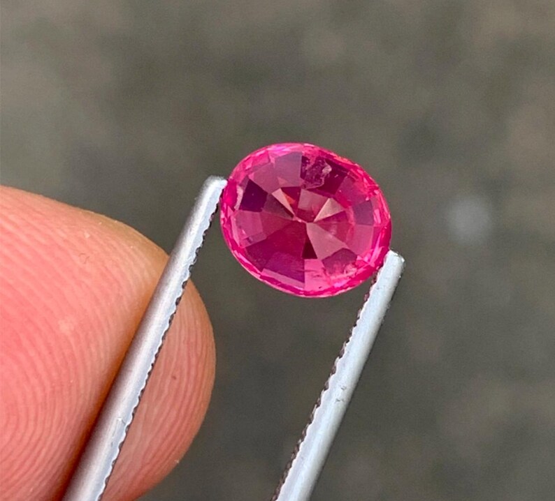May include: A faceted, oval-cut, hot pink gemstone is held by silver tweezers. The gem has a brilliant cut, reflecting light. The background is a blurred, neutral tone.