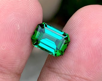 May include: A vibrant green emerald gemstone cut in an emerald shape. The gemstone is held between two fingers.