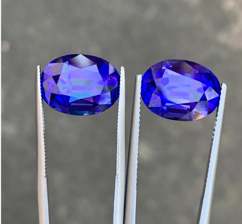 May include: Two oval-shaped gemstones, both a deep purple-blue color, are held by tweezers against a gray background.