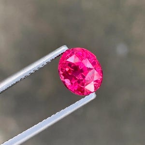 May include: A faceted, oval-cut, hot pink gemstone is held by a pair of silver tweezers. The gem has many facets that reflect light, creating a brilliant sparkle. The background is a blurred, neutral tone.