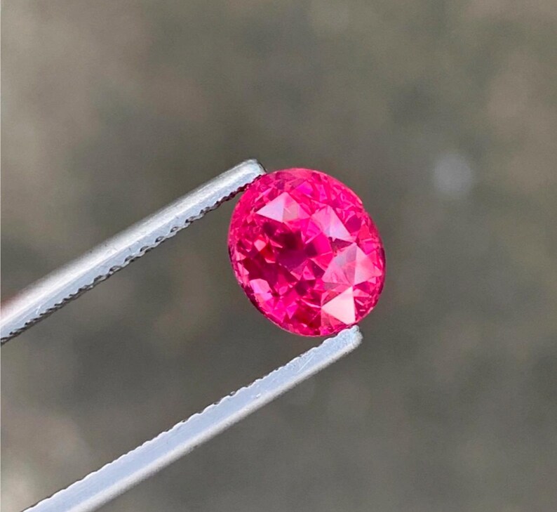 May include: A faceted, oval-cut, hot pink gemstone is held by a pair of silver tweezers. The gem has many facets that reflect light, creating a brilliant sparkle. The background is a blurred, neutral tone.