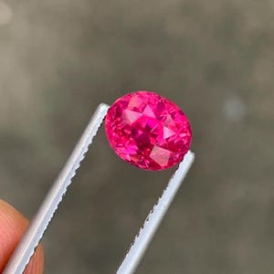 May include: A faceted, oval-cut, hot pink gemstone is held by silver tweezers against a blurred gray background. The gem's many facets reflect light, creating a sparkling effect. The tweezers are held by a person's fingers.