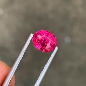 May include: A faceted, oval-shaped, hot pink gemstone is held by silver tweezers. The gem has a brilliant cut, reflecting light. The background is a blurred, neutral tone.
