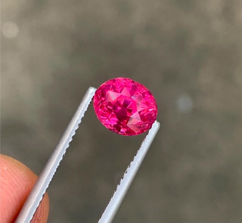 May include: A faceted, oval-shaped, hot pink gemstone is held by silver tweezers. The gem has a brilliant cut, reflecting light. The background is a blurred, neutral tone.
