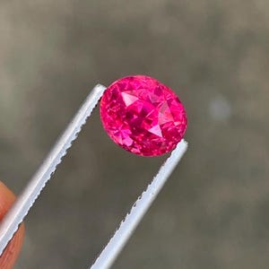 May include: A faceted, oval-shaped, hot pink gemstone is held by a pair of tweezers. The gem has a brilliant cut, reflecting light. The background is a blurred, neutral gray.