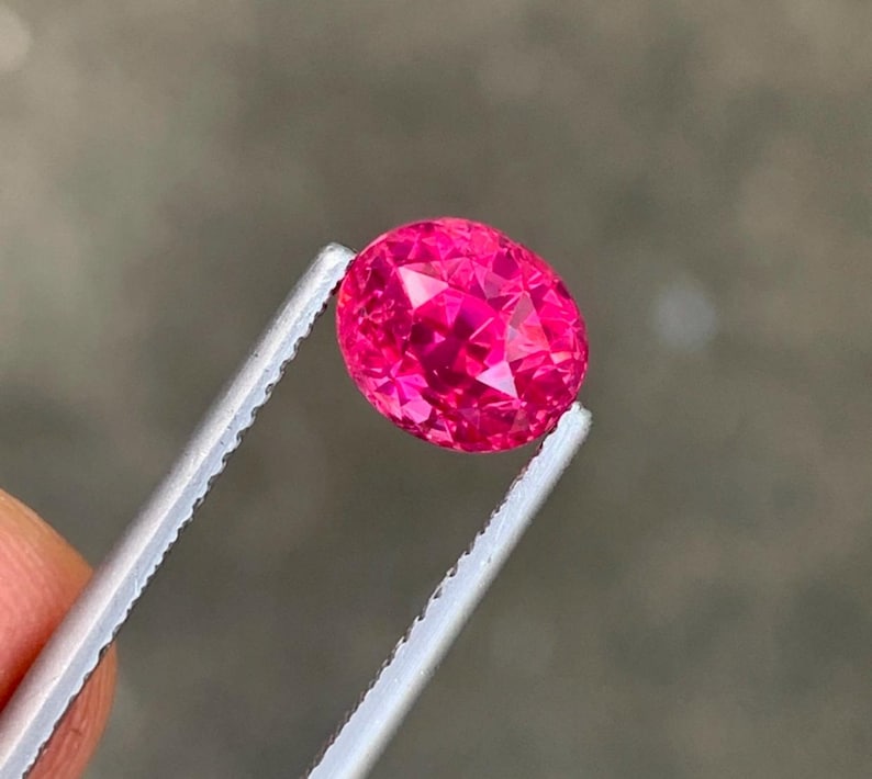 May include: A faceted, oval-shaped, hot pink gemstone is held by a pair of tweezers. The gem has a brilliant cut, reflecting light. The background is a blurred, neutral gray.