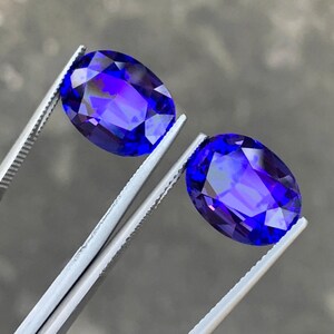 May include: Two oval-shaped, deep blue gemstones with a purple hue, held by silver tweezers.