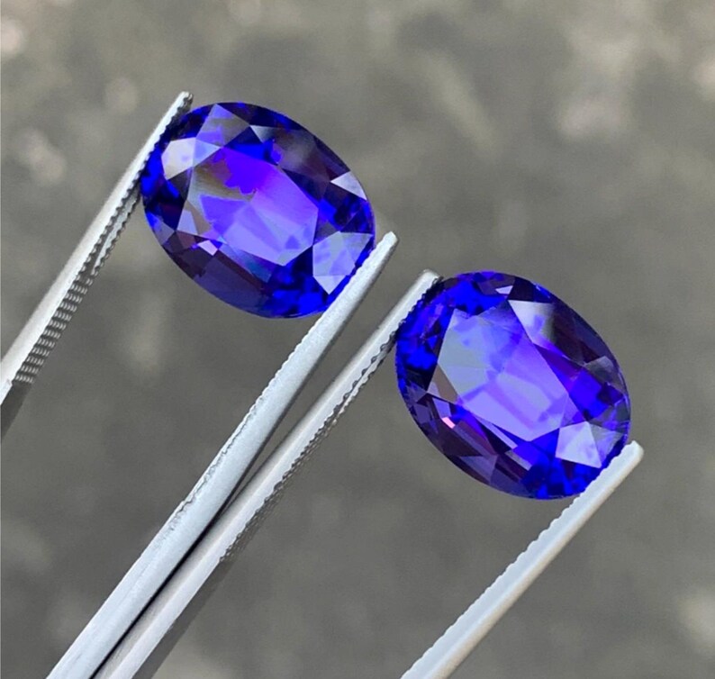 May include: Two oval-shaped, deep blue gemstones with a purple hue, held by silver tweezers.