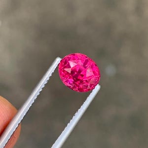 May include: A faceted, oval-shaped, hot pink gemstone is held by a pair of silver tweezers. The gemstone has a brilliant cut, reflecting light. The tweezers are held by a person's fingers.