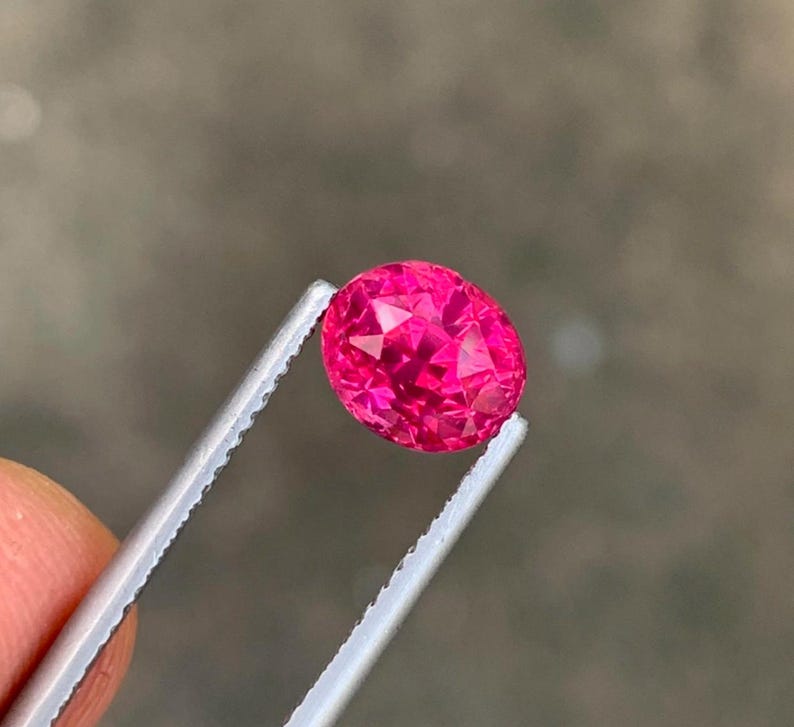 May include: A faceted, oval-shaped, hot pink gemstone is held by a pair of silver tweezers. The gemstone has a brilliant cut, reflecting light. The tweezers are held by a person's fingers.