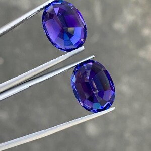 May include: Two oval-shaped, faceted gemstones with a deep blue to purple color. The gemstones are held by tweezers.