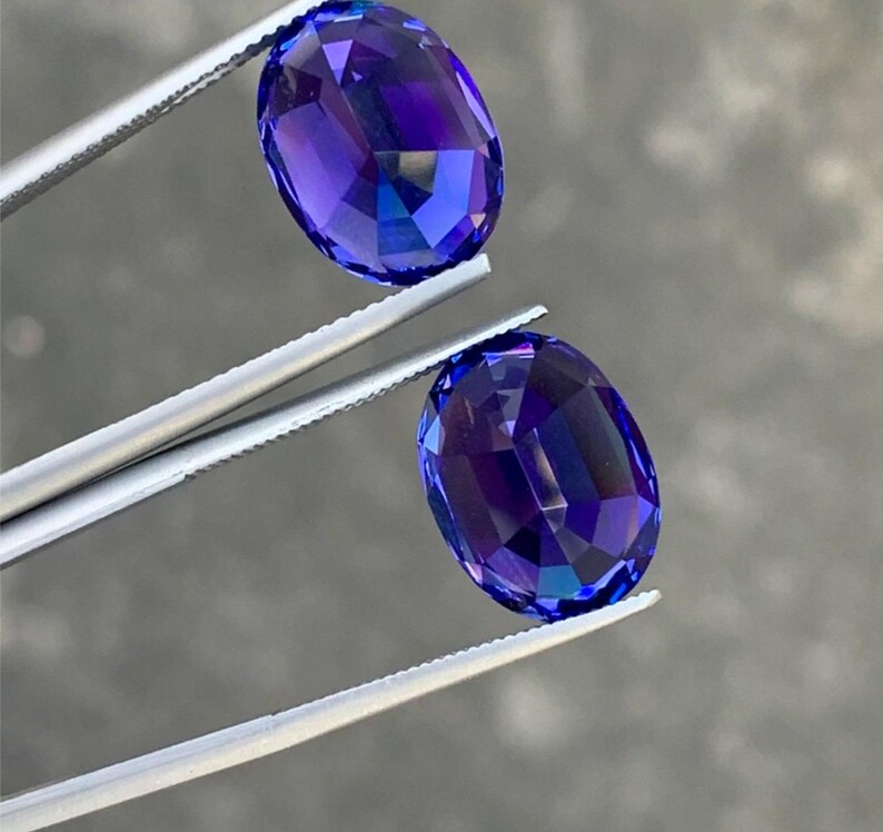 May include: Two oval-shaped, faceted gemstones with a deep blue to purple color. The gemstones are held by tweezers.
