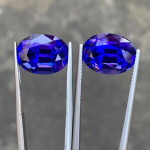 May include: Two oval-shaped, deep blue gemstones held by tweezers. The gemstones are faceted and have a high level of brilliance.