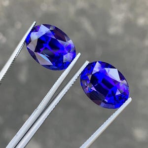 May include: Two oval-shaped, deep blue gemstones held by tweezers. The gemstones are faceted and have a high polish.