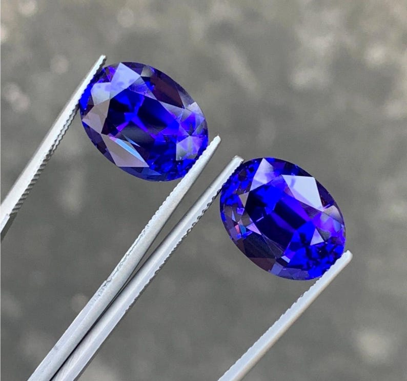 May include: Two oval-shaped, deep blue gemstones held by tweezers. The gemstones are faceted and have a high polish.