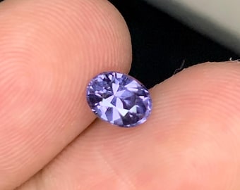 May include: An oval-shaped, faceted, lavender-blue gemstone. The gem is held between two fingers, showcasing its polished surface and internal reflections. The stone's color is a vibrant, medium tone, and it appears to be a high-quality precious stone.