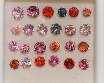 May include: A collection of 20 faceted gemstones in a white display case. The gemstones are arranged in rows and are various shades of pink, red, and orange.