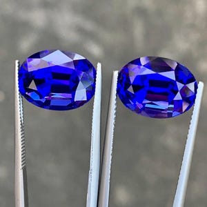 May include: Two oval-shaped, deep blue gemstones are held by tweezers. The gemstones are faceted and have a high polish.