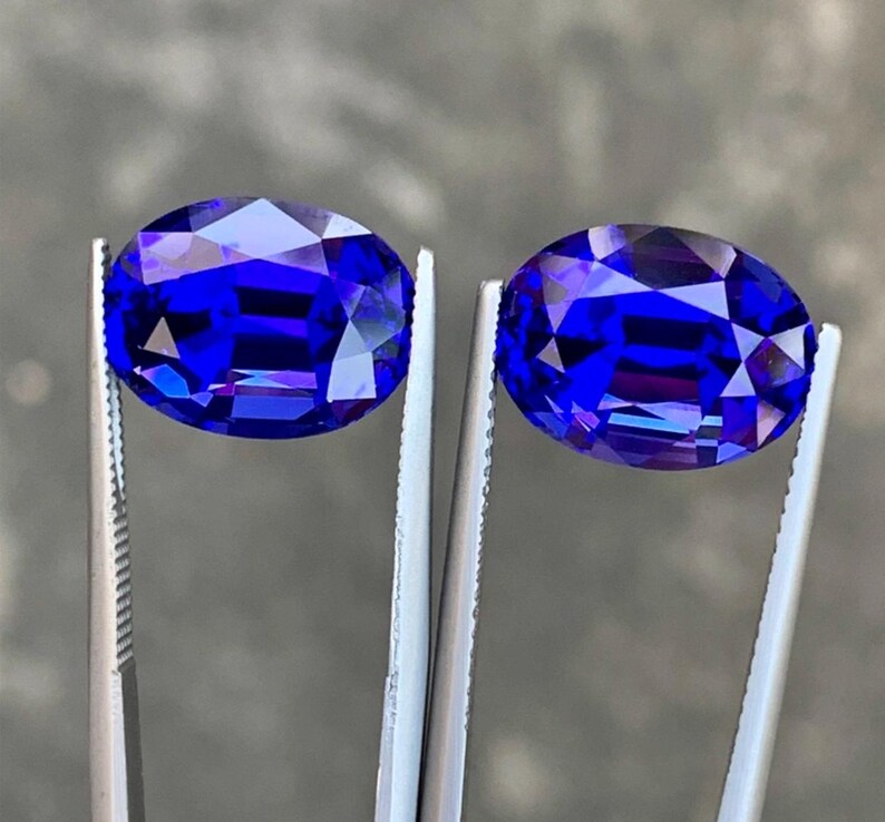 May include: Two oval-shaped, deep blue gemstones are held by tweezers. The gemstones are faceted and have a high polish.