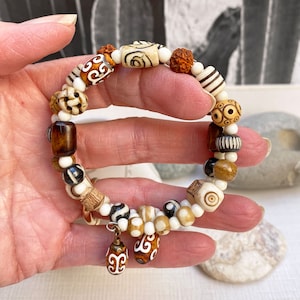 May include: A bracelet made of white and brown beads with intricate patterns. The bracelet is made of various materials, including bone, wood, and stone. The bracelet is worn on a person's wrist.