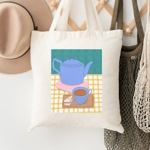 Könnte beinhalten: Eine weiße Stofftasche mit einer blauen Teekanne, einer rosa-weißen Teetasse und einem Keks auf einer gelb-weißen karierten Tischdecke. Die Stofftasche hängt an einem Holzgestell mit einem braunen Hut und einer beigefarbenen Netztasche.