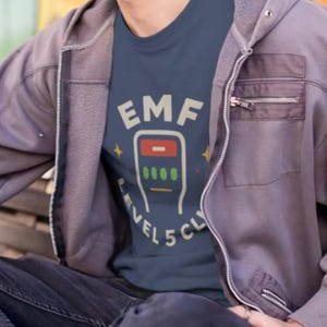 May include: A navy blue t-shirt with the text "EMF Levels 5 CLY" in white. The graphic includes a red and green design. The person is wearing a gray zip-up hoodie and dark pants.