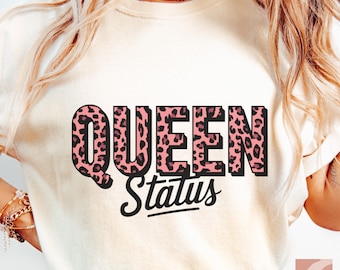 May include: Cream-colored t-shirt with the words "QUEEN Status" in a black and pink leopard print design. The word "QUEEN" is in a bold, blocky font, and "Status" is in a script font.