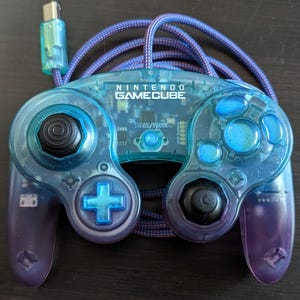 May include: A transparent blue Nintendo GameCube controller with a purple cable. The controller has black joysticks and buttons, with the text "NINTENDO GAMECUBE" visible on the top. The cable is a braided purple and blue design.